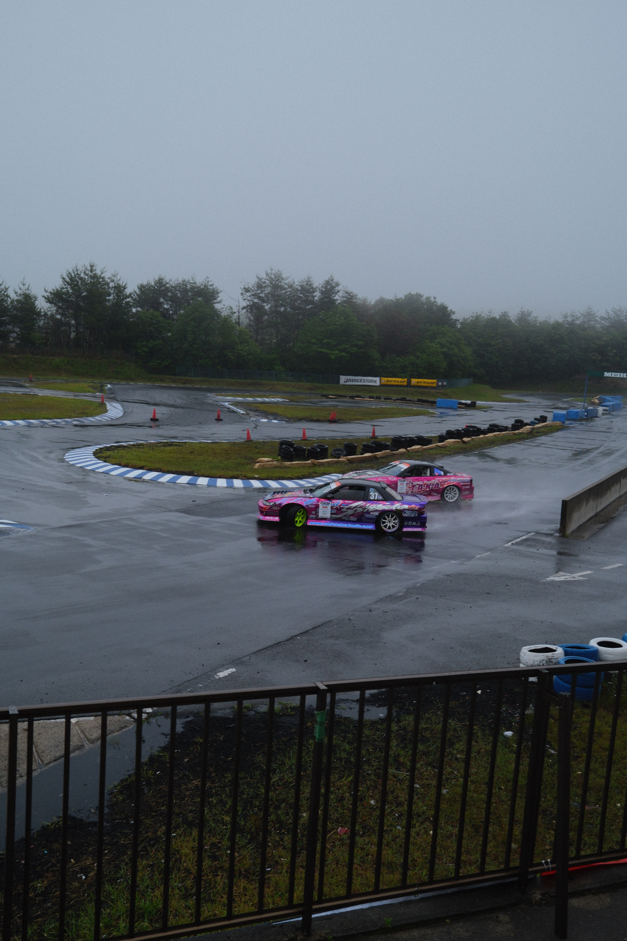 Two drift cars sliding through a hairpin in heavy rain and mist